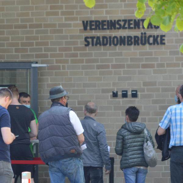 Menschen stehen in einer Schlange vor einem Gebäude mit einem Schild mit der Aufschrift Vereinszentrum Stadionbrücke.