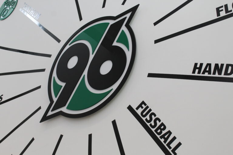 Nahaufnahme des Logos von Hannover 96 mit schwarzen Linien und deutschen Wörtern, die von ihm ausgehen.