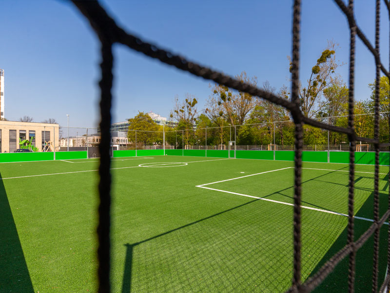 Blick auf einen leeren Fußballplatz mit grünem Kunstrasen, gesehen durch ein schwarzes Netz unter freiem Himmel.