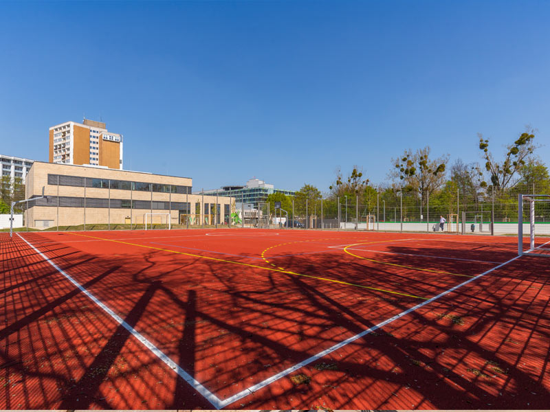 Ein roter Sportplatz im Freien mit weißen und gelben Linien, Gebäuden und Bäumen im Hintergrund unter einem klaren Himmel.