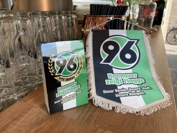 Hannover 96 Wimpel und Karte auf einer Theke mit Gläsern und Strohhalmen im Hintergrund.