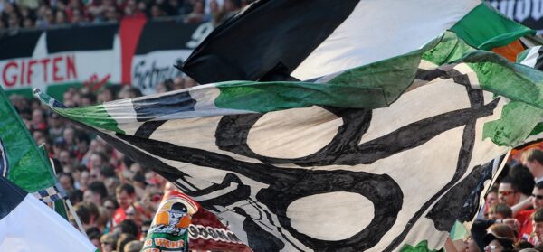 Eine große Menschenmenge in einem Stadion schwenkt große schwarz-weiß-grüne Fahnen mit auffälligen Motiven.
