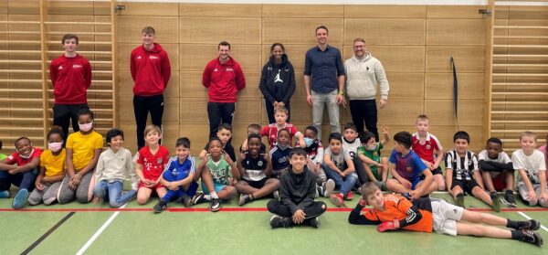 Eine Gruppe von Kindern und Erwachsenen posiert gemeinsam in einer Turnhalle, einige sitzen, andere stehen und lächeln in die Kamera.