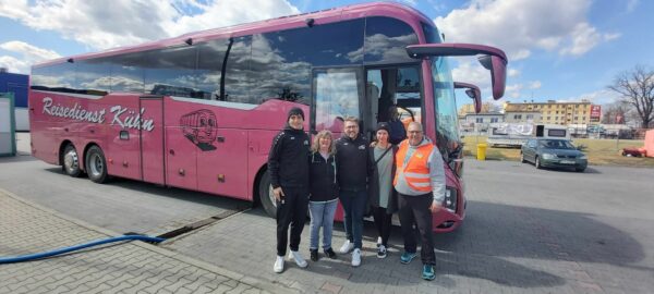 Fünf Personen stehen lächelnd vor einem großen rosafarbenen Tourbus auf einem gepflasterten Parkplatz unter einem teilweise bewölkten Himmel.
