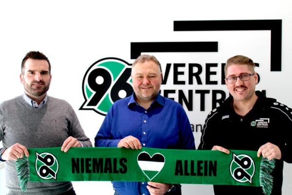 Drei Männer halten einen grünen Schal mit der Aufschrift Niemals Allein vor einem Hannover 96-Logo an einer Wand.
