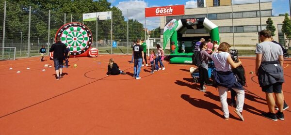 Auf einem Außenplatz werden Spiele angeboten, darunter eine riesige Dartscheibe und eine aufblasbare Hüpfburg.