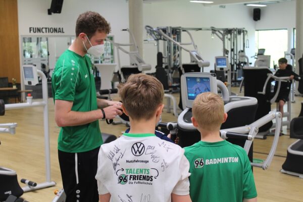 Ein Trainer mit Maske instruiert zwei Jungen in Sporttrikots an Fitnessgeräten in einer Turnhalle.