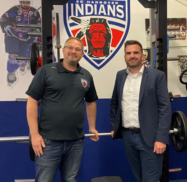 Zwei Männer stehen vor einem EC Hannover Indians-Logo in einer Turnhalle, an der Wand sind Eishockeyfotos zu sehen.