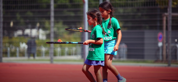 Zwei junge Mädchen in grünen Hemden spielen Tennis mit Schlägern und orangefarbenen Bällen auf einem Platz im Freien.