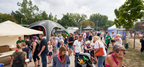 Eine Menschenmenge versammelt sich unter freiem Himmel bei einem Festival mit Zelten, Ständen und Bäumen unter einem bewölkten Himmel.