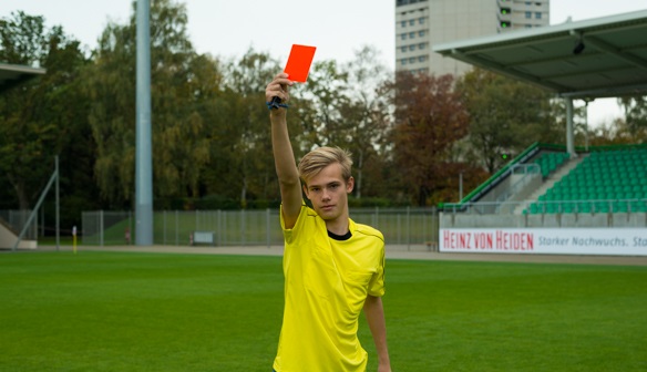 Fußballschiedsrichter in gelbem Hemd hält eine rote Karte auf einem grünen Feld mit leeren Tribünen im Hintergrund hoch.